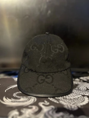 Svart Gucci keps med GG-mönster - Snygg svart keps från Gucci med det klassiska GG-monogrammet över hela kepsen. Kepsen har böjd skärm och justerbar rem bak. Tillverkad i ett slitstarkt tygmaterial som ger en exklusiv känsla. Perfekt för dig som vill ha en stilren accessoar med lyxig touch.