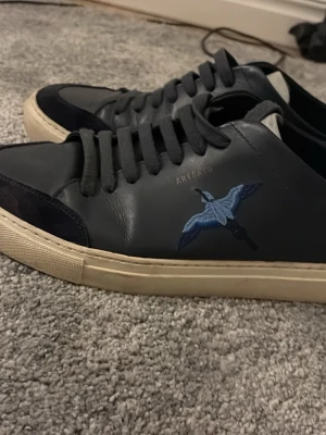 Svarta Axel Arigato sneakers med broderi - Snygga svarta sneakers från Axel Arigato med blått broderat fågelmotiv på sidan. Skorna har svarta skosnören, svart mockadetalj vid tån och vit sula. Tillverkade i läder och mocka för en clean och modern look.