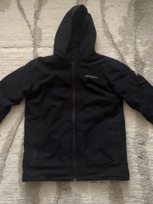 Svart vindjacka från Peak Performance - Snygg svart vindjacka från Peak Performance med huva och hel dragkedja framtill. Jackan har två sidofickor med dragkedja och en diskret logga på bröstet. Tillverkad i ett lätt och tåligt material som skyddar mot vind.