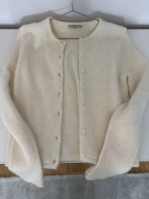 Krämvit kofta från Gina Tricot  - Mysig krämvit kofta från Gina Tricot med rund halsringning och knäppning framtill. Tillverkad i ett mjukt, fluffigt material som ger en avslappnad och stilren look. Perfekt att slänga över en t-shirt eller topp för extra värme.