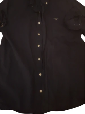 Mörkblå skjorta från GANT - Klassisk mörkblå skjorta från GANT med knappstängning framtill och broderad logga på bröstet. Skjortan har lång ärm och en tidlös design som passar till mycket. Tillverkad i mjuk bomull för skön känsla.