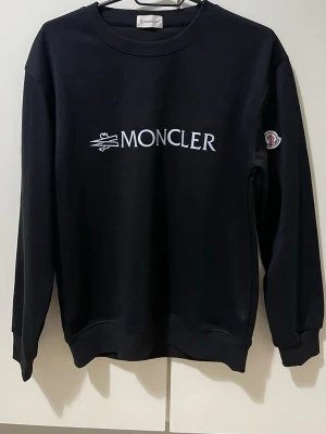Svart Moncler sweatshirt - Snygg svart sweatshirt från Moncler med vit logga tryckt på bröstet och ett broderat Moncler-märke på ärmen. Tröjan har rund halsringning, ribbade muddar vid ärmslut och nederkant. Perfekt för en stilren och trendig look.