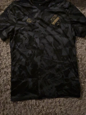 Svart AIK x Nike matchtröja - Säljer en svart AIK matchtröja från Nike med guldfärgade loggor på bröstet. Tröjan har ett diskret kamouflagemönster och är tillverkad i ett lätt, snabbtorkande material som passar perfekt till träning eller match.