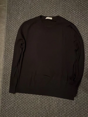 Svart stickad tröja Tiger of Sweden - Klassisk svart stickad tröja från Tiger of Sweden. Tröjan har rund halsringning, långa ärmar och en stilren, minimalistisk design. Perfekt basplagg i garderoben och enkel att matcha. Materialet ser ut att vara mjukt och följsamt.