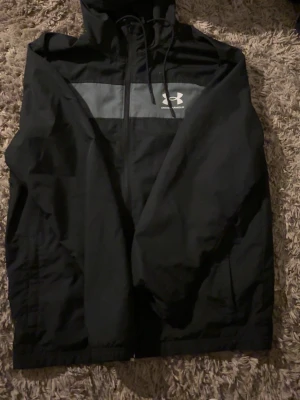 Svart vindjacka från Under Armour - Snygg svart vindjacka från Under Armour med dragkedja framtill och huva med dragsko. Jackan har en grå panel över bröstet med Under Armour-loggan tryckt i vitt. Perfekt för blåsiga dagar och sportiga outfits.