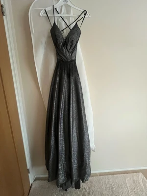 Glittrig långklänning med snörning - Snygg långklänning i glittrigt mörkgrått tyg med tunna axelband och korsad snörning i ryggen. Klänningen har v-ringad urringning och ett elegant fall. Perfekt för bal eller festkvällar när du vill sticka ut. Orginal pris 4000