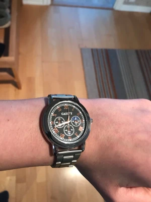 Svart-silver klocka - Stilren klocka från Gaiety med svart urtavla, romerska siffror i guld och tre dekorativa subdials. Boetten och länken är i silverfärgad metall vilket ger en klassisk look. Perfekt accessoar för dig som gillar tidlös design.