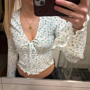 Vit topp med blå blommor och volang - Söt vit topp med små blå blommor, volangkant och knytning framtill. Djup v-ringning och långa ärmar med lätt utsvängda avslut. Stretchigt ribbat material som sitter tajt och framhäver figuren. Perfekt för en trendig och ungdomlig stil.