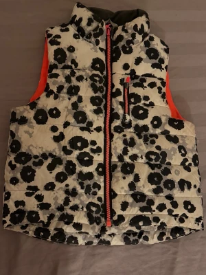 Svartvit dunväst med leopardmönster - Cool dunväst med svart och grått leopardmönster på vit botten och orange detaljer vid dragkedjan och sidorna. Hög krage och en bröstficka med dragkedja. Perfekt för dig som vill sticka ut och hålla värmen under hösten eller vintern.