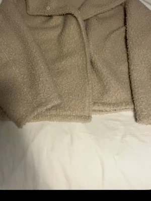 Beige cropped teddyjacka - Mysig beige teddyjacka i kort modell med bred krage och mjukt, fluffigt material. Jackan har långa ärmar och stängs med knappar framtill. Perfekt för dig som vill ha en trendig och bekväm look under kyliga dagar.