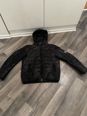 Moncler jacka herr svart polyamid storlek 50 - Snygg svart moncler jacka med huva och dragkedja framtill. Jackan har två fickor med dragkedja och quiltad design. Perfekt för kalla dagar och har en stilren look. Märke syns på ärmen men är otydligt. Materialet är glansigt och lätt vadderat.