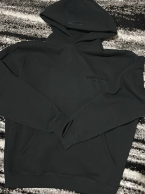 Svart Essentials Fear of God hoodie - Säljer en svart hoodie från Fear of God Essentials med diskret logga på bröstet och stor text på ryggen. Hoodien har huva, magficka och ribbade muddar. Perfekt för dig som gillar streetwear och vill ha en clean look.
