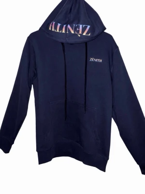 Mörkblå hoodie från Zénith - Snygg mörkblå hoodie från Zénith med tryckt logga på bröstet och i regnbågsfärger på huvan. Klassisk känguruficka framtill, justerbar dragsko i huvan och mjukt material som känns skönt mot huden. Perfekt för en chill och avslappnad stil. 