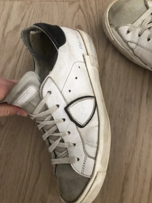 Vita Philippe Model sneakers - Snygga vita sneakers från Philippe Model med grå mockadetaljer på tån och svart hälflik. Skorna har en ikonisk sköldlogga på sidan och vita skosnören. Tillverkade i läder och mocka för en trendig och avslappnad look.