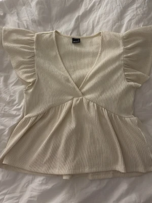 Krämvit med volangärm - Supersöt krämvit topp med v-ringning och volangärmar. Toppen har en lätt ribbad struktur och ett flowy fall nedtill som ger en söt vibe. Perfekt till jeans eller kjol för en trendig look. kontakta för fler bilder på toppen!!