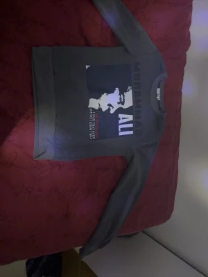 Grå stickad tröja Muhammad Ali - Grå sweatshirt med stort tryck av Muhammad Ali och citat på framsidan. Tröjan har rund hals, långa ärmar och ribbade muddar. Materialet ser ut att vara mjukt och bekvämt, perfekt för en sportig och avslappnad stil.