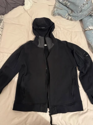 Svart hoodie från C.P. Company - Säljer en svart hoodie från C.P. Company med dragkedja framtill och huva. Hoodien har en ficka med dragkedja på ena ärmen och klassisk logodetalj. Tillverkad i mjukt och bekvämt material, perfekt för en avslappnad streetwear-look.