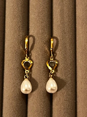 Guldörhängen med pärldroppar - Snygga örhängen i guldfärgad metall med hjärtformad detalj och vita droppformade pärlor. Örhängena har en klassisk klickstängning och en elegant design som ger en lyxig känsla. Perfekta för dig som gillar stilrena accessoarer med det lilla extra.