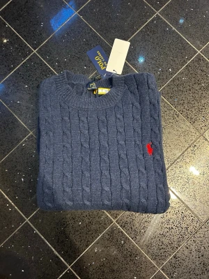 Blå kabelstickad tröja Polo Ralph Lauren - Kabelstickad blå tröja från Polo Ralph Lauren med rund halsringning och den klassiska röda logotypen broderad på bröstet. Tröjan har långa ärmar och ribbade muddar vid ärmslut och nederkant. Perfekt för dig som gillar stilren och tidlös design.