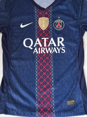 PSG matchtröja blå Nike - Snygg blå matchtröja från Paris Saint-Germain med röd och vit detalj i mitten, FIFA-märke och klubbmärke på bröstet. Tillverkad i lätt och ventilerande material, perfekt för fotboll eller träning. Qatar Airways-logga på framsidan.