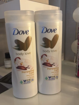 Dove Body Love Pampering Care lotion - Två flaskor Dove Body Love Pampering Care body lotion med sheasmör och vaniljdoft. Flaskorna är vita med guld- och bronsfärgade detaljer och har en elegant, rundad form. Perfekt för återfuktning och mjuk hud.