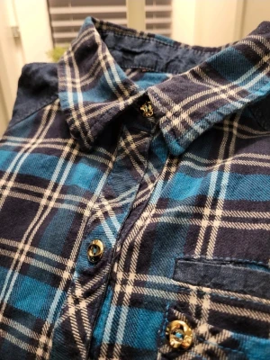 Blå rutig skjorta med guldknappar S - Snygg blå och svart rutig skjorta med vita detaljer och guldiga knappar. Skjortan har klassisk krage, bröstficka och långa ärmar. Perfekt för en avslappnad men trendig look.