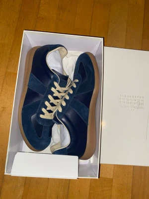 Maison Margiela blå sneakers - Snygga sneakers från Maison Margiela i mörkblå mocka och läder med beigea skosnören och gummisula. Klassisk low-top modell med stilren design och ikoniska detaljer. Perfekta för dig som vill ha något unikt och trendigt i garderoben.