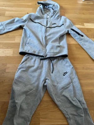 Gråa mjukisbyxor från Nike  - Snygga gråa mjukisbyxor från Nike med svart logga på vänster ben. Byxorna har dragsko i midjan och sidofickor. Tillverkade i mjukt och bekvämt material, perfekta för chill eller träning. Matchar med hoodie för en komplett look.