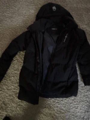 Svart dunjacka från Sail Racing - Säljer en svart dunjacka från Sail Racing med huva och dragkedja. Jackan är vindtät, andas och har DWR-behandling samt certifierat dun. Märket syns tydligt på bröstet och insidan. Perfekt för kalla vinterdagar och har en sportig look.