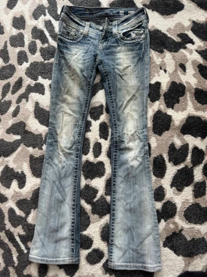 Blå bootcut jeans från Miss Me - Säljer nu tyvärr mina favorit jeans från Miss me då de blivit alldeles för små för mig. Det är ett par blå/grå lågmidjade bootcut jeans från Miss Me med slitna detaljer och dekorativa nitar vid fickorna. Skit snygga jeans med paljett detaljer på fickorna och baksidan i lila & silver. Storlek 23, perfekt i längd på mig som är ca 162 cm☺️Jag har köpt dom av en tjej här på plick, jeansen är absolut använda en hel del gånger men inga större defekter eller slitningar🙌