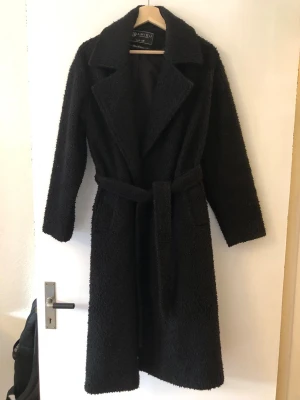 Svart lång teddyjacka med skärp - Säljer en svart teddyjacka från Jaimisu med lång passform och breda slag. Jackan har ett matchande midjebälte och två fickor framtill. Perfekt för dig som vill ha en mysig och trendig look under kalla dagar.