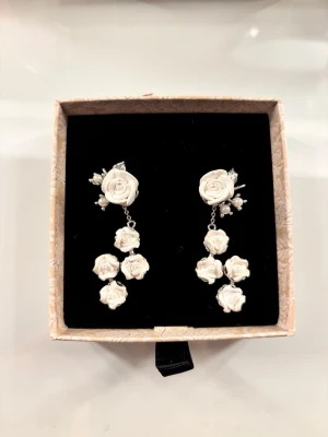 Örhängen från ioaku - Unika örhängen med vita rosor i flera nivåer och små pärldetaljer. Örhängena har en elegant och romantisk design med blommor i plast eller keramik och silverfärgade metalldelar. 