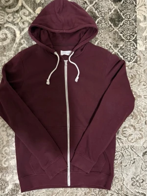 Pier one Hoodie - Snygg vinröd hoodie från Pier One med dragkedja framtill och vita dragsnören i huvan. Tillverkad i mjukt bomullsmaterial och har två fickor fram. Perfekt för en avslappnad och stilren look.