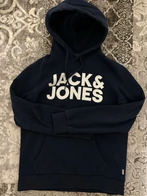 Mörkblå hoodie från Jack & Jones - Snygg mörkblå hoodie från Jack & Jones med stor vit logga på bröstet. Klassisk känguruficka framtill och justerbar huva med dragsko. Tillverkad i mjukt bomullsmaterial som är skönt mot huden. Perfekt för en avslappnad och trendig look.