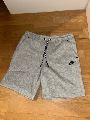Grå  Nike tech schorts med snörning - Grå sweatshorts från Nike med svart logga på vänster ben och snörning i midjan. NIKE TECH SCHORTS, pris kan diskuteras, skriv för mer bilder 