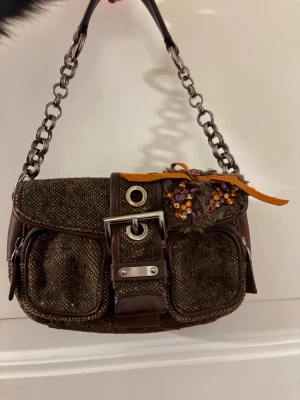 Brun handväska från Prada med kedja - Unik brun handväska från Prada i grovt tyg och läderdetaljer. Väskan har en dekorativ kedjerem, två fickor framtill, spänne och broderade blommor med pärlor och orange band. Perfekt accessoar för dig som gillar detaljer och exklusiv design.