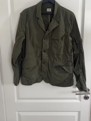 Olivgrön kavaj från C.P. Company - Stilren olivgrön kavaj från C.P. Company med tre framfickor och knappstängning. Tillverkad i lätt, skrynkligt material som ger en avslappnad look. Klassisk krage och enkel design, perfekt för dig som gillar streetwear med en twist. I storlek Medium och inköpt på United Fashion i göteborg. Jag väger 77kg och är 182cm. Letar efter ett gött bud så tveka inte på att skriva!