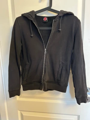 Svart hoodie från Colmar - En svart hoodie med dragkedja från Conrad. Hoodien har huva med dragsko, två fickor framtill och ribbade muddar vid ärmar och nederkant. Tillverkad i mjukt bomullsmaterial som är skönt att ha på sig. Perfekt för en avslappnad och enkel stil.