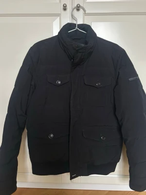 Svart dunjacka från Woolrich - Snygg svart dunjacka från Woolrich med hög krage och fyra framfickor med lock och knapp. Klassisk och stilren design med diskret logga på ärmen. Perfekt för kalla vinterdagar tack vare det varma dunfodret.