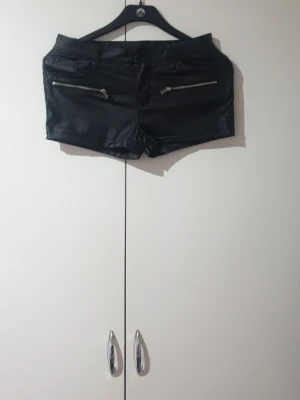 Svarta shorts i skinnimitation - Snygga svarta shorts i skinnimitation med dragkedjedetaljer framtill och fickor både fram och bak. De har en midwaist passform och ger en edgy vibe till din outfit. Perfekta för dig som vill sticka ut lite extra.