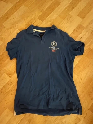 Blå piké t-shirt från Henri Lloyd - Klassisk blå pikétröja från Henri Lloyd med korta ärmar och broderad logga samt brittisk flagga på bröstet. Tröjan har krage och knappar vid halsen. Perfekt för en avslappnad och stilren look.