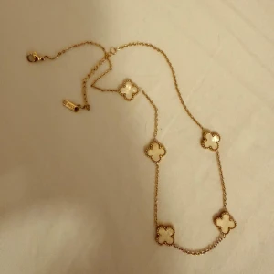 Guldhalsband med vita fyrklöver - Snyggt halsband i guldton med fem vita fyrklöverformade hängen. Kedjan är tunn och elegant, och varje hänge har en pärlliknande kant. Perfekt för dig som gillar stilrena och trendiga accessoarer.