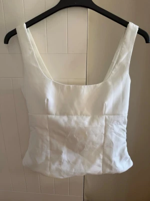 Vit linnetopp med rosett i ryggen - Stilren vit linnetopp med breda axelband och fyrkantig ringning. Toppen har en snygg rosettdetalj i ryggen som ger en extra touch. Perfekt att matcha med jeans eller kjol för en fräsch look. Materialet känns svalt och lätt. Köpt på plick innan och har inget märke eller lapp för storlek i sig. Jag skulle säga att de är en S/M