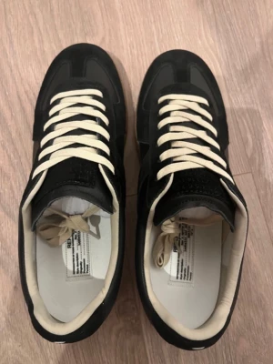  Maison Margiela gats  - Maison Margiela Replica sneakers i svart mocka och läder med vita kontrasterande skosnören och beige innersula. Klassisk låg modell med gummisula och diskret logotyp på plösen. Perfekta för dig som gillar stilrena och tidlösa sneakers.