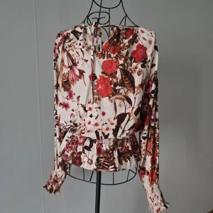 Supersöt blus med blommigt mönster – rött, brunt och beige  - En feminin blus med vackert blommigt mönster i varma toner. Vida, långa ärmar med smock vid handlederna och charmig volang nertill som ger en lekfull touch. Rund halsringning med knyt i nacken och ett flowy, luftigt fall som känns både elegant och bekvämt. Perfekt till vardag eller festligare tillfällen ✨