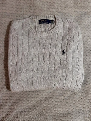 Ralph Lauren - Kabelstickad tröja i grå från Polo Ralph Lauren med rund halsringning och klassisk broderad logga på bröstet. Tröjan har ribbade muddar vid ärmslut och nederkant och är tillverkad i ett mjukt material som passar perfekt till höst och vinter. Längd 61cm & bredd 40 cm 