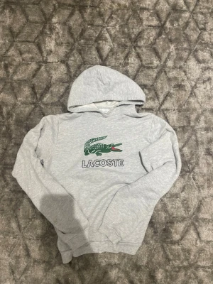 Grå hoodie från Lacoste med logga - Snygg ljusgrå hoodie från Lacoste med den klassiska gröna krokodilloggan och texten 'LACOSTE' på bröstet. Hoodien har huva och är tillverkad i mjukt bomullsmaterial. Perfekt för en chill och sportig look. Skickas inom 24h.