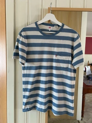 Randig Levi's t-shirt i blått och vitt - Säljer en randig t-shirt från Levi's i blått och vitt med bröstficka och klassisk rund hals. Skön passform och tillverkad i mjuk bomull. Perfekt för dig som gillar en avslappnad och stilren look. Stl S 