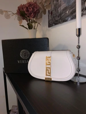 Vit axelremsväska från Versace - Säljer en snygg vit handväska från Versace med guldfärgade detaljer och ett elegant geometriskt spänne framtill. Väskan har en rundad form och exklusiv känsla, perfekt för dig som vill sticka ut med en lyxig accessoar.
