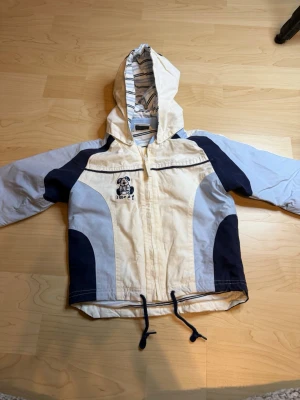 Vit och blå vindjacka KappAhl Baby - Söt vindjacka från KappAhl Baby i vitt och marinblått med ljusblå detaljer. Jackan har huva med randigt foder, dragkedja framtill, dragsko i nederkant och ett gulligt hundmotiv på bröstet. Perfekt för blåsiga dagar och lek utomhus.